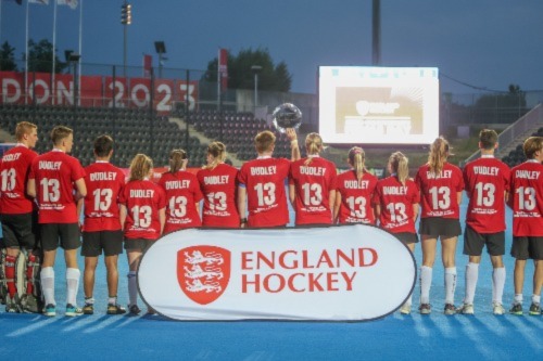 Bristol hockey players stand with their back to the camera. On their red shirts are the name 'Dudley' and number '13'. They are standing on a blue hockey pitch with a sign that says 'England Hockey'.